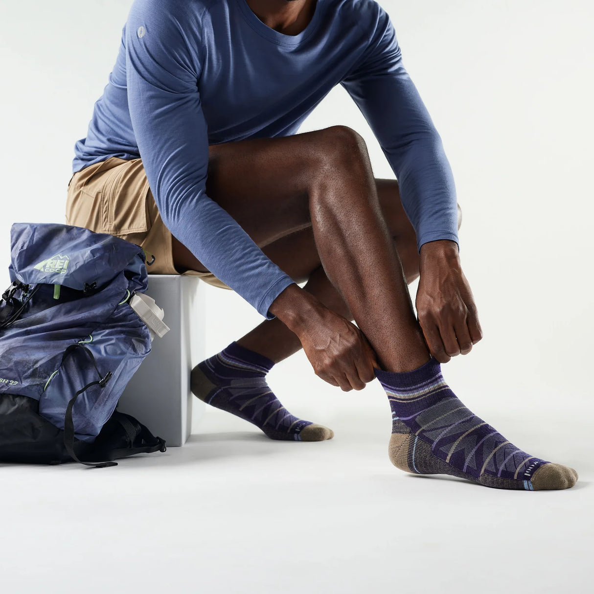 Person wearing blue long-sleeve shirt and patterned socks, sitting with a backpack on a white background - Smartwool Hike Pattern Light Cushion Ankle Socks (Unisex) - Nightfall Blue