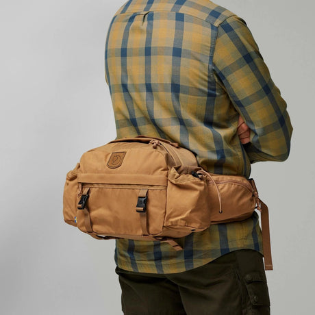 Person wearing a tan waist bag with a plaid shirt and dark pants on a gray background - Fjällräven Singi Hip Pack 10 - Khaki Dust