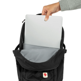 A black Fjälör laptop backpack shown open with a hand inserting a silver laptop into the top compartment. The backpack features a gray interior lining and a small red and white Fjälör logo patch on the front pocket, set against a plain white background. - Fjällräven Skule 28 Backpack - Black