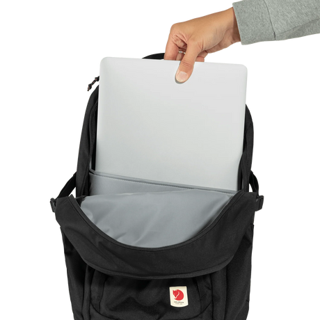 A black Fjälör laptop backpack shown open with a hand inserting a silver laptop into the top compartment. The backpack features a gray interior lining and a small red and white Fjälör logo patch on the front pocket, set against a plain white background. - Fjällräven Skule 28 Backpack - Black