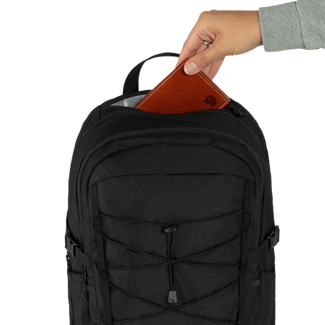 A black backpack shown from the front with a hand inserting a brown leather wallet into the top zipper compartment, against a plain white background. - Fjällräven Skule 28 Backpack - Black