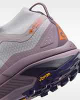 A Nike ACG ZEGAMA ZOOMX trail running shoe in a light lavender and purple colorway, featuring a textured mesh upper, a purple Swoosh, and a purple Vibram outsole, shown from a side profile on a light gray background.