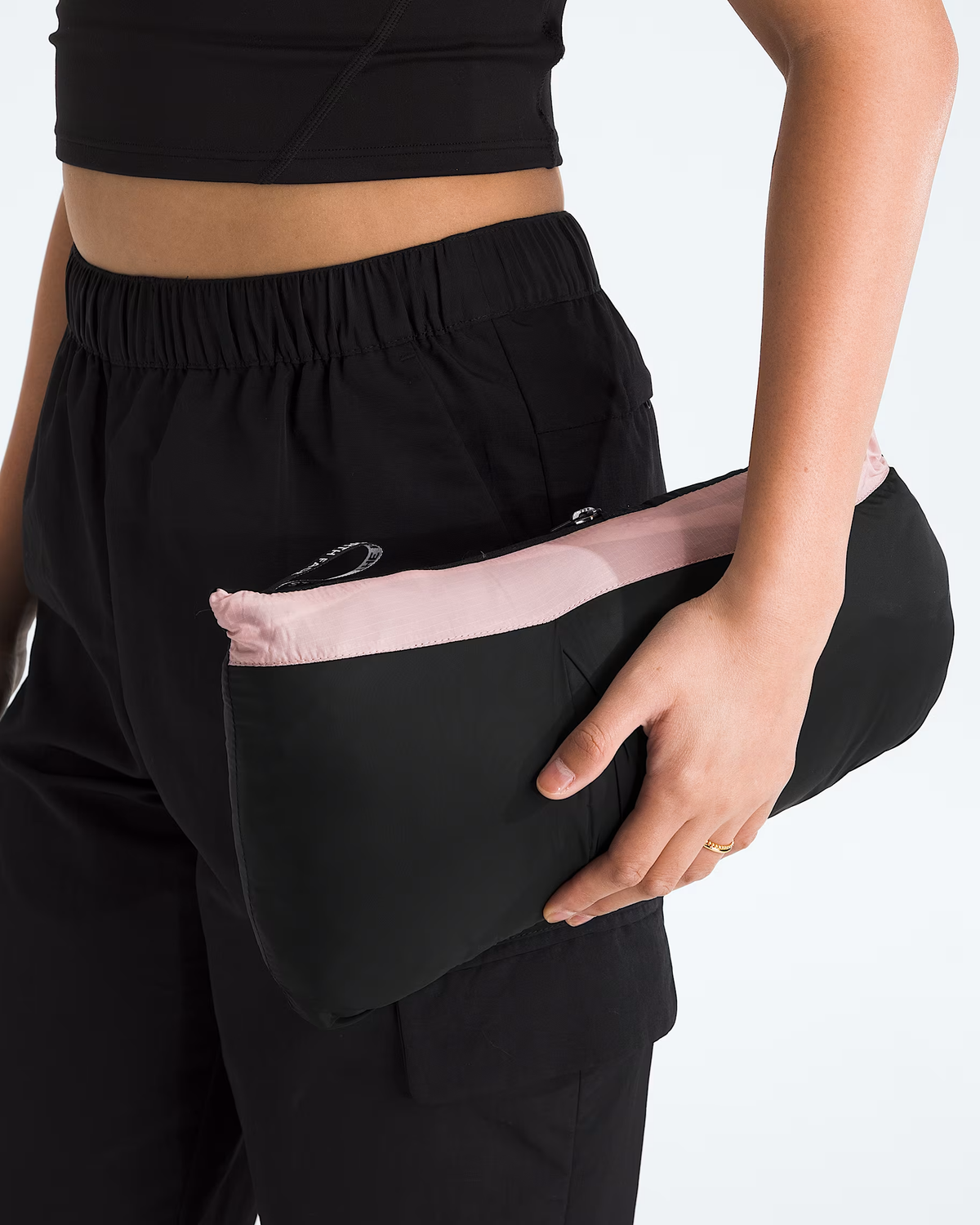 A close-up of a person wearing black athletic pants and holding a black pouch with a light pink interior lining.- The North Face 1996 Retro Nuptse Jacket (Womens) - Metal Pink/TNF Black