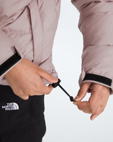 Close-up of a person wearing a light pink puffer jacket and black pants, adjusting the drawstring of the jacket. The North Face logo is visible on the thigh.- The North Face 1996 Retro Nuptse Jacket (Womens) - Metal Pink/TNF Black