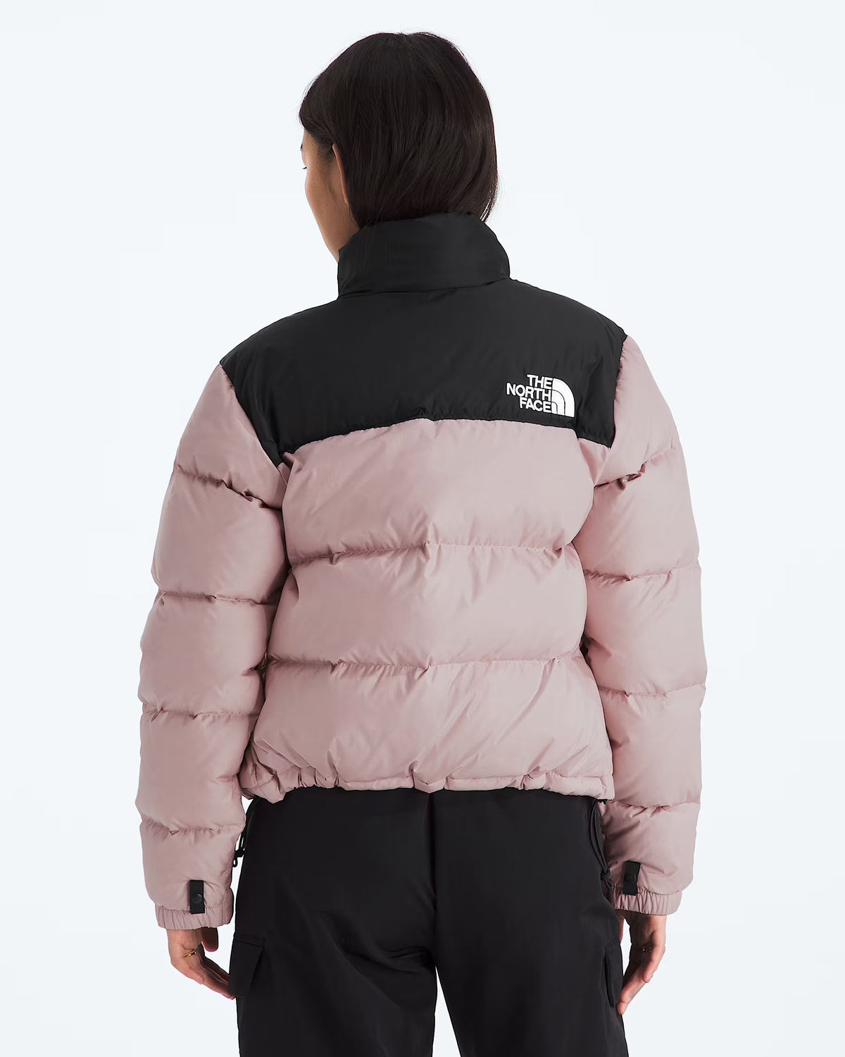 A model viewed from the back wearing a colorblock puffer jacket by The North Face. The jacket features a black upper yoke and collar, and a dusty pink body with horizontal quilting.- The North Face 1996 Retro Nuptse Jacket (Womens) - Metal Pink/TNF Black