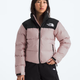 A woman wearing a light pink and black puffer jacket from The North Face, paired with black pants, against a plain light gray background.- The North Face 1996 Retro Nuptse Jacket (Womens) - Metal Pink/TNF Black