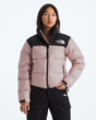 A woman wearing a light pink and black puffer jacket from The North Face, paired with black pants, against a plain light gray background.- The North Face 1996 Retro Nuptse Jacket (Womens) - Metal Pink/TNF Black