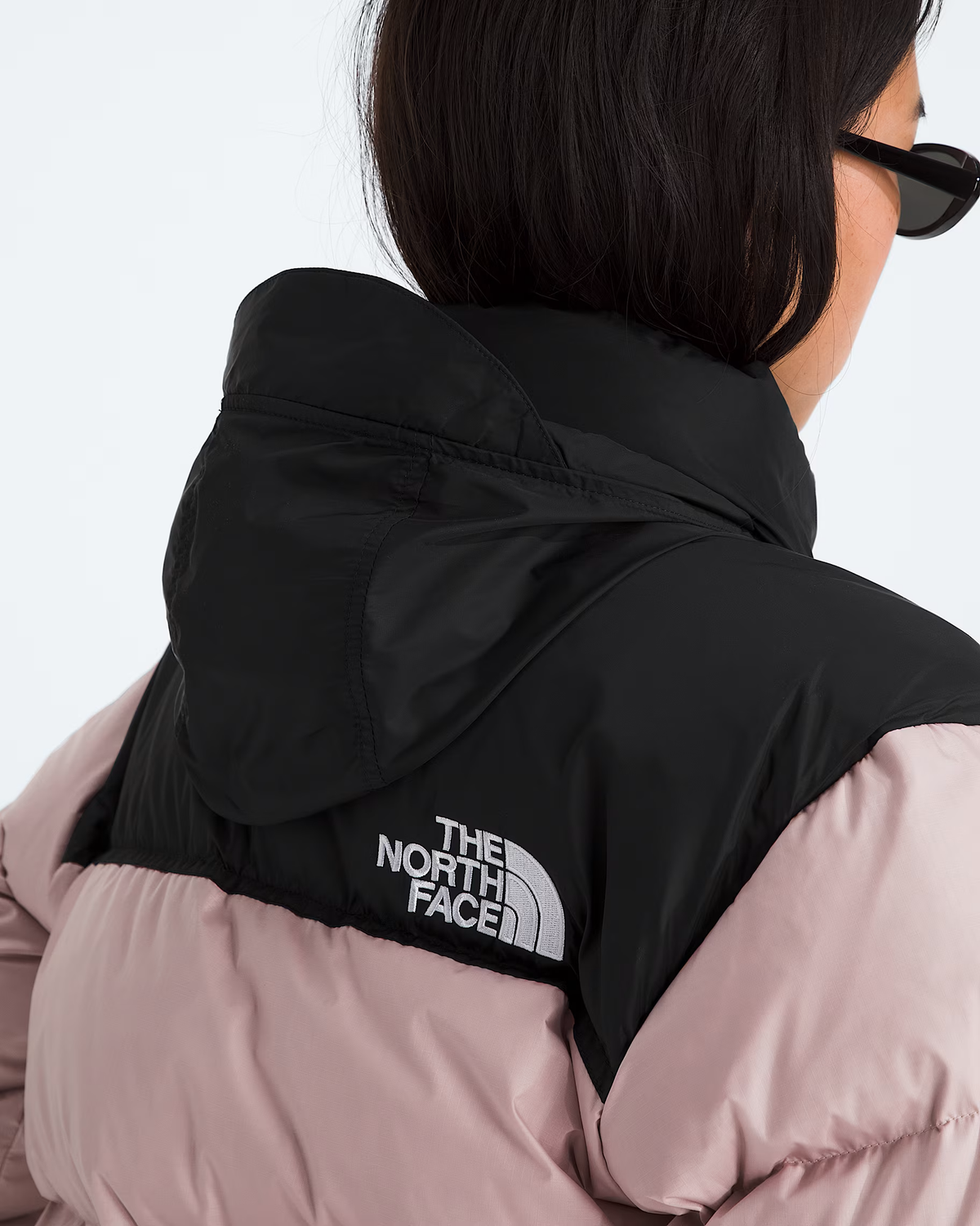 Close-up back view of a person wearing a black hooded puffer vest over a pale pink puffer jacket, with The North Face logo visible on the back.- The North Face 1996 Retro Nuptse Jacket (Womens) - Metal Pink/TNF Black