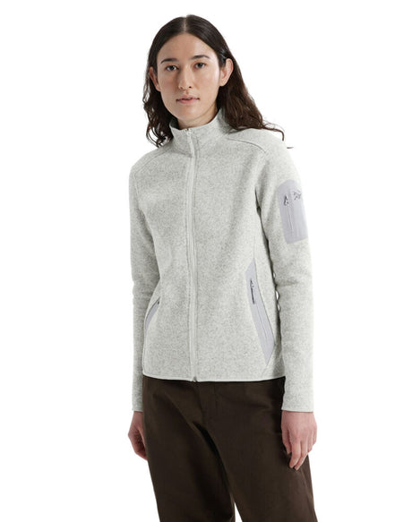 A woman modeling a light gray heather Arc'teryx zip-up fleece jacket with a stand-up collar, zippered pockets, and a light gray logo patch on the upper sleeve, paired with dark brown pants against a plain white background. - Arc'teryx Covert Cardigan (Women's) - Atmos Heather
