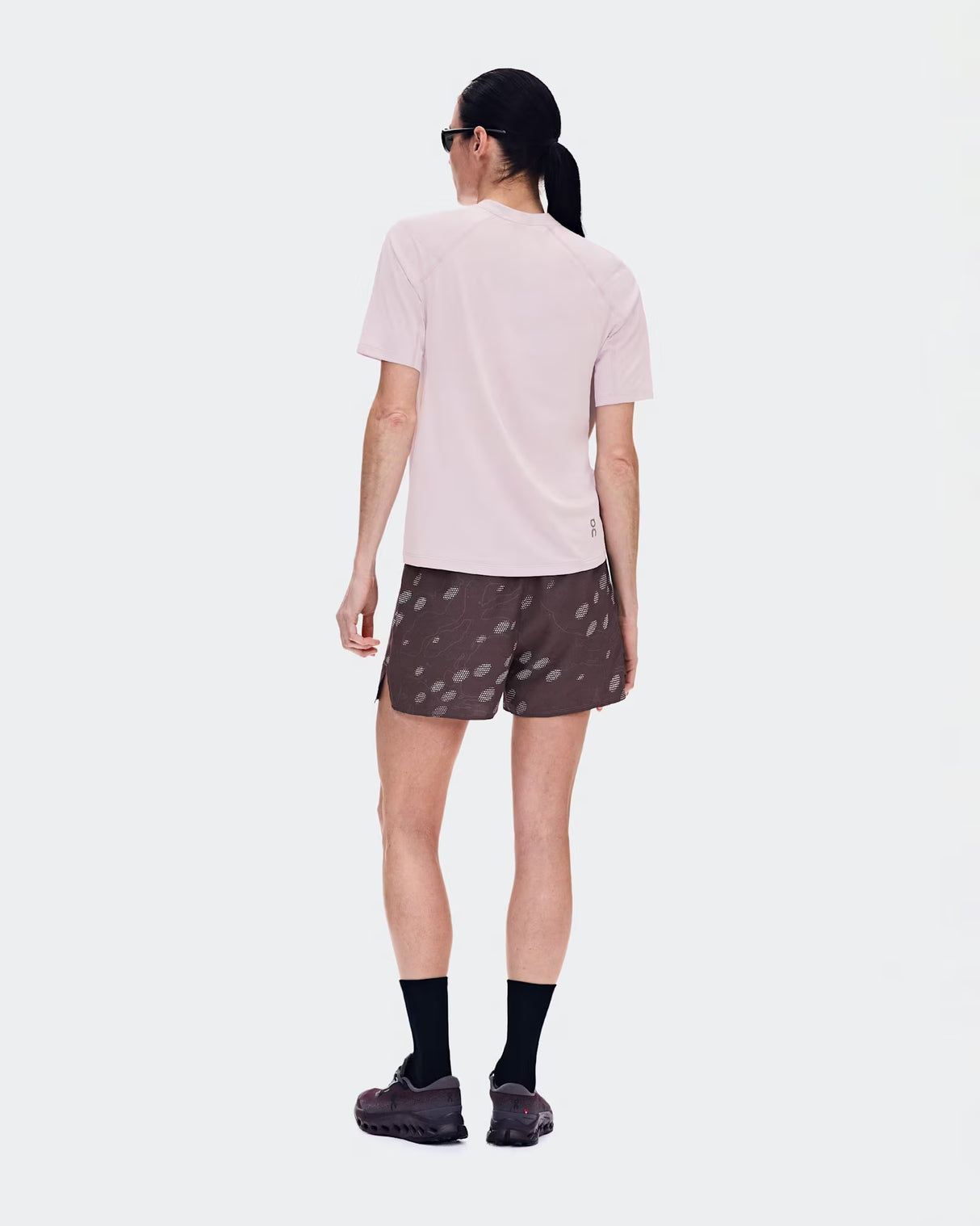 A woman is shown from behind wearing a light pink short-sleeve athletic t-shirt, dark brown patterned running shorts, black socks, and trail running shoes against a light gray studio background. - On Trail Shorts (Women's) - Mulberry