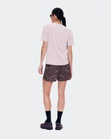 A woman is shown from behind wearing a light pink short-sleeve athletic t-shirt, dark brown patterned running shorts, black socks, and trail running shoes against a light gray studio background. - On Trail Shorts (Women's) - Mulberry