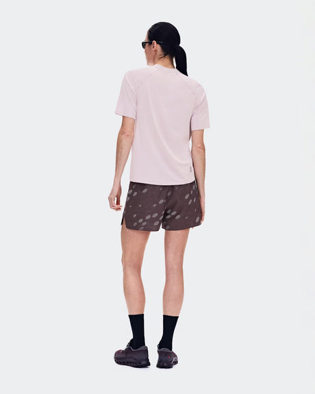 A woman is shown from behind wearing a light pink short-sleeve athletic t-shirt, dark brown patterned running shorts, black socks, and trail running shoes against a light gray studio background. - On Trail Shorts (Women's) - Mulberry