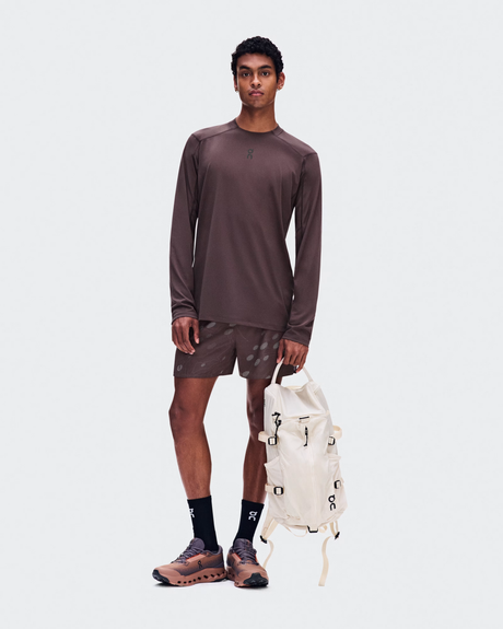 A male model stands against a light gray studio background wearing a matching brown athletic long-sleeve shirt and shorts set, black socks, and trail running shoes, while holding a cream-colored backpack. - On Trail Long-T Solar (Men's) - Mulberry
