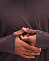 Close-up of a person wearing a dark brown athletic long-sleeve top with the On logo near the collar, showing the elasticized wrist opening and hands stretching the sleeve. - On Trail Long-T Solar (Men's) - Mulberry
