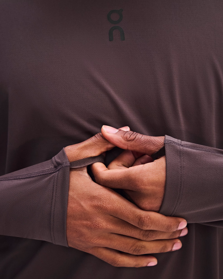 Close-up of a person wearing a dark brown athletic long-sleeve top with the On logo near the collar, showing the elasticized wrist opening and hands stretching the sleeve. - On Trail Long-T Solar (Men's) - Mulberry