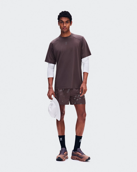 A male model stands against a light gray studio background wearing a dark brown athletic t-shirt layered over a white long-sleeve shirt, matching brown patterned shorts, black socks, and brown running shoes, while holding a white cap. - On Trail-T Solar (Men's) - Mulberry
