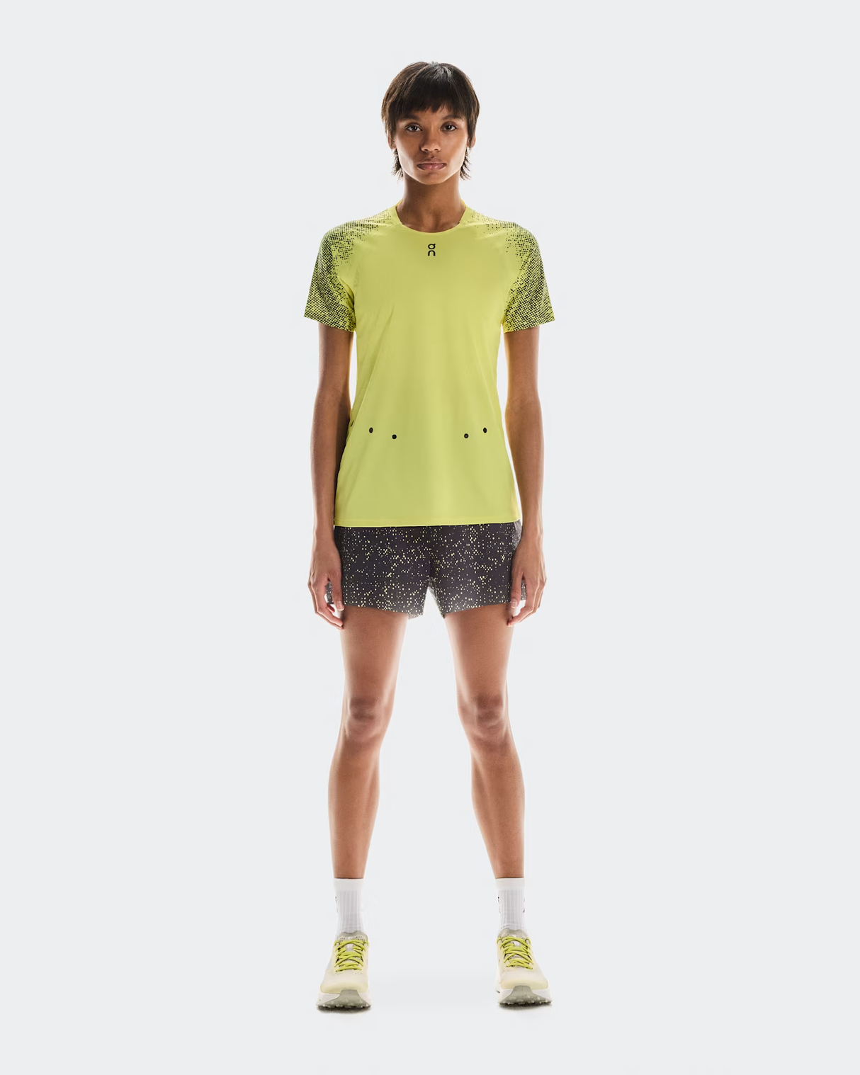 Person wearing a bright yellow athletic shirt and black patterned shorts on a white background - On Ultra Shorts (Women's) - Iron/Lime