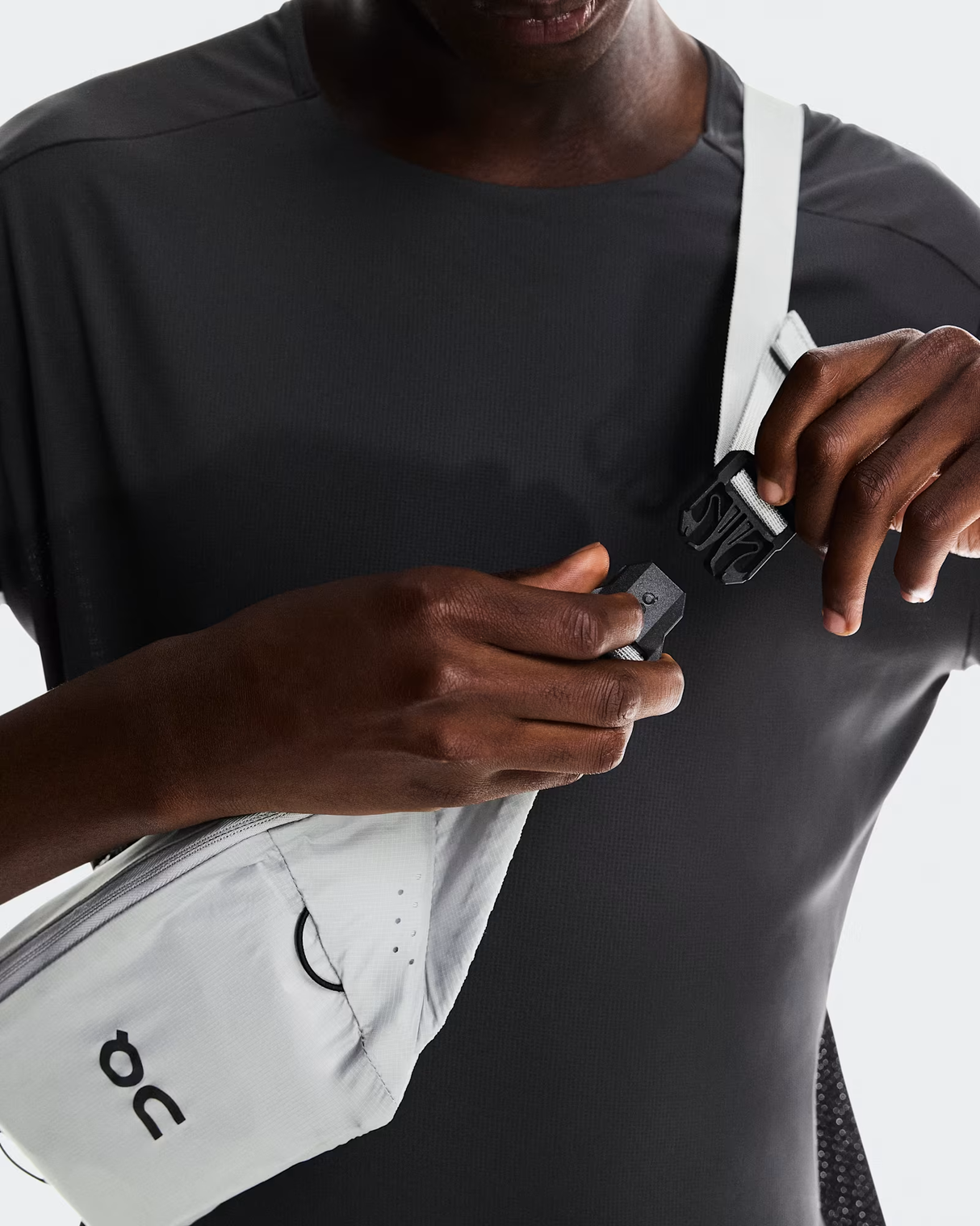 Person adjusting a white bag strap with a visible brand logo on a plain background - On Waist Pack 2L Lite - Glacier