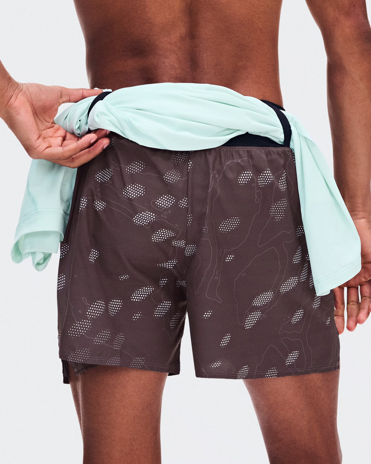 A close-up back view of a model wearing dark brown swim trunks with a white topographic pattern, with a mint green shirt tied around the waist. - On Trail Shorts (Men's) - Mulberry
