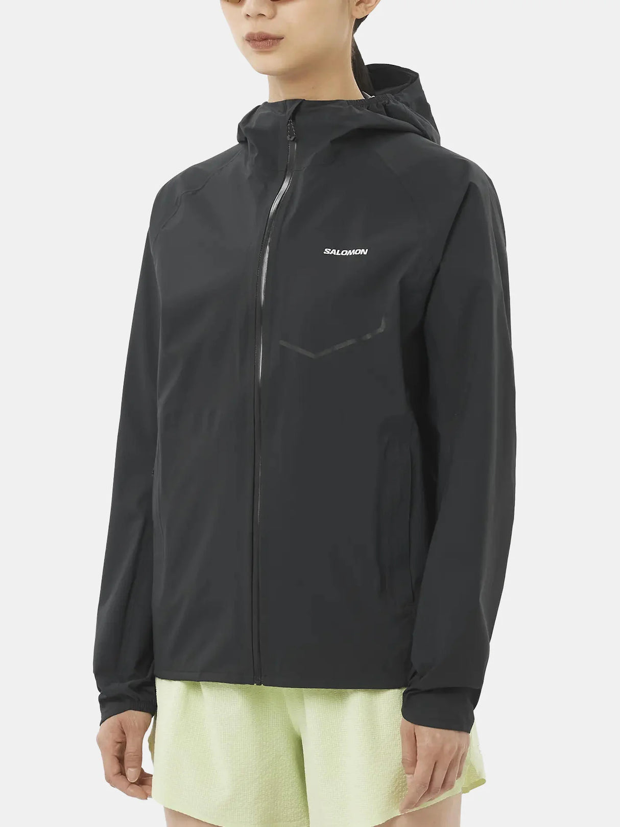 A model wearing a black hooded athletic jacket with a full front zipper and a small white MULLION logo on the left chest, paired with light green shorts, against a plain light gray background. - Salomon Bonatti Waterproof Jacket (Women's) - Deep Black