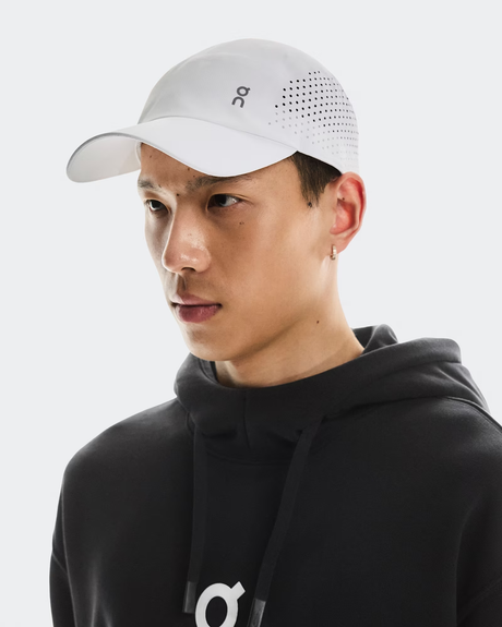 Person wearing a black hoodie and white cap with a logo on a light gray background - On Lightweight Cap - White